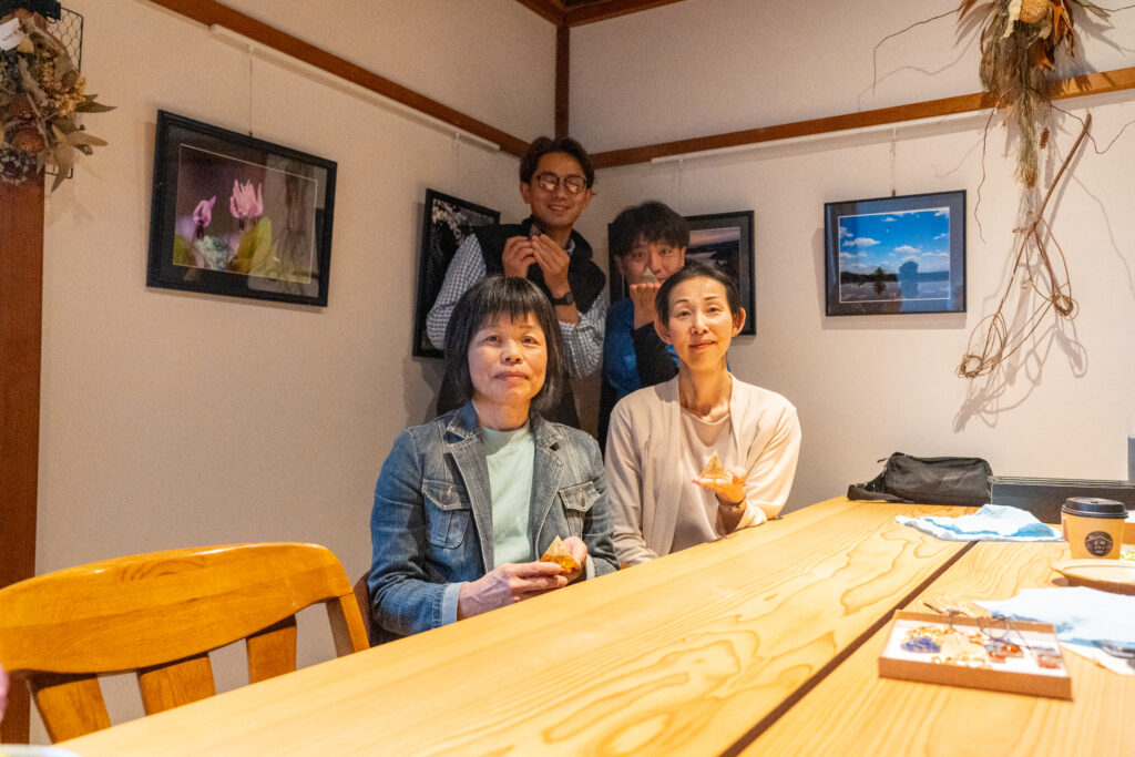 「上町コテジで行われた第5回スマホ撮影ワークショップの様子。参加者3名がカフェのテーブルを囲み、焼き菓子を手に持ちながら楽しそうにポーズをとっている。背景には花や風景の写真が飾られている。」