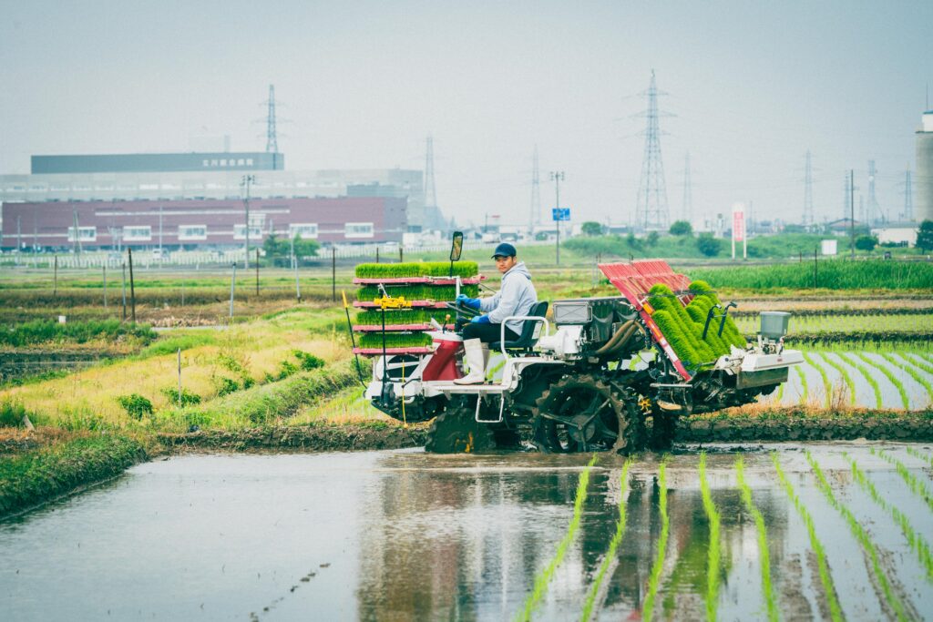 田植え機に乗って作業中の農家の男性スタッフ。背景には長岡市の工場と送電塔、水田に広がる苗の列が整然と映る。