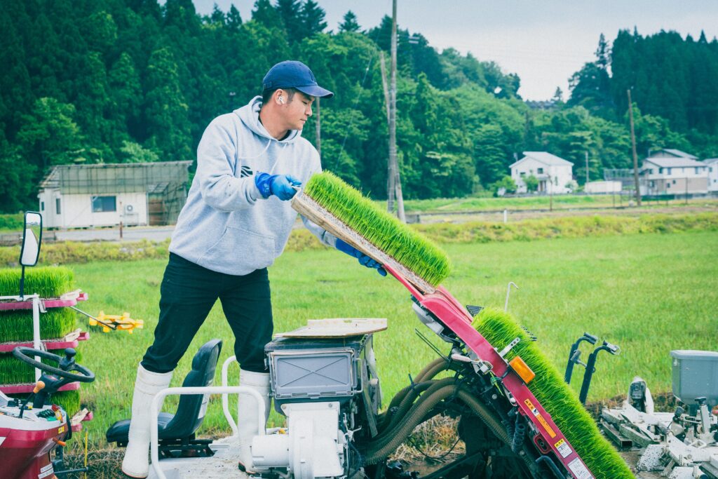 農作業中の男性スタッフが、田植え機に苗箱をセットしている様子。背景には木々と住宅が並ぶ、緑豊かな風景が広がる。