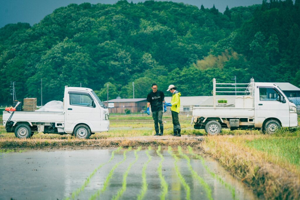 水田のあぜ道で話すふたりのスタッフ。両脇には苗や資材を載せた軽トラックが停まり、作業の合間の打ち合わせ風景をとらえた写真。