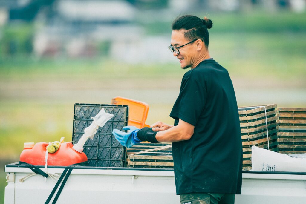 軽トラックの荷台で準備作業をする男性スタッフ。苗箱や給油タンクなどが積まれた車両のそばで、笑顔を浮かべながら手袋をはめている。