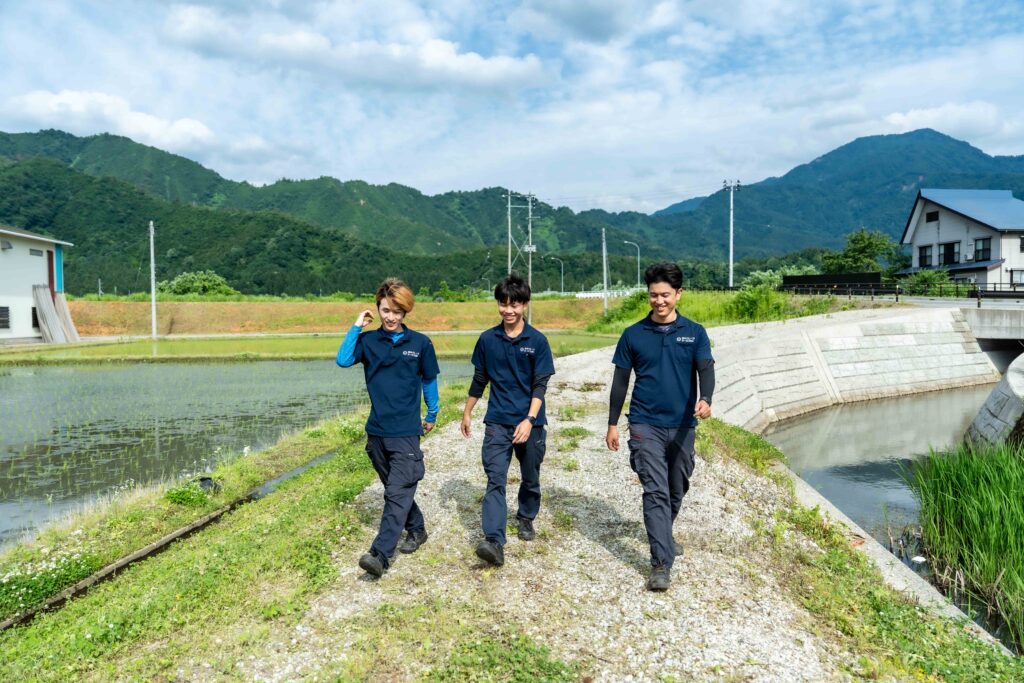 南魚沼の美しい山々と田園風景をバックに、笑顔で言葉を交わしながら歩く3人の若手社員。