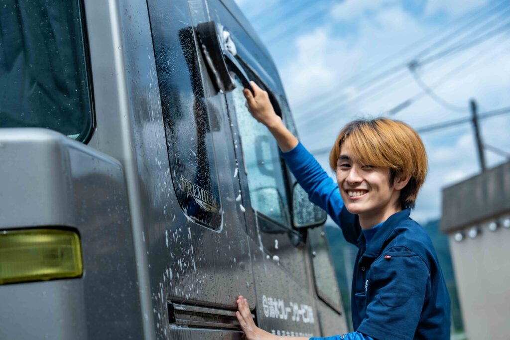 笑顔でクレーン車の窓ガラスを磨き上げる若手社員。道具を大切にするプロ意識と明るい現場の雰囲気。